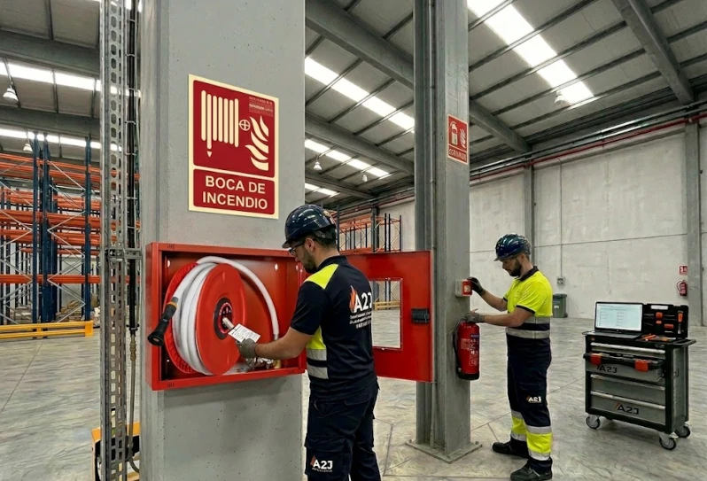 Técnicos de A2J Mantenimientos Integrales realizando la revisión de una Boca de Incendio Equipada (BIE) y un extintor portátil en una nave logística de Sevilla. Se observa el uso de un carro de herramientas digitalizado y software de control para garantizar el cumplimiento del RIPCI.